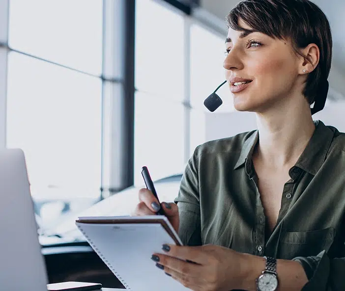 Frau mit Headset sitzt an einem Schreibtisch, hält einen Notizblock und spricht.