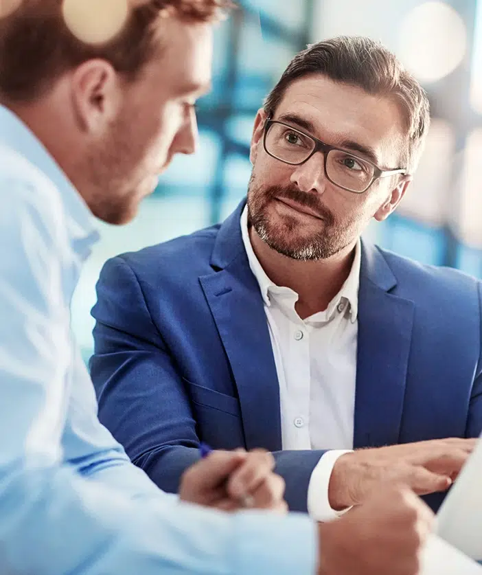 Zwei Männer in Businesskleidung führen ein Gespräch am Schreibtisch.